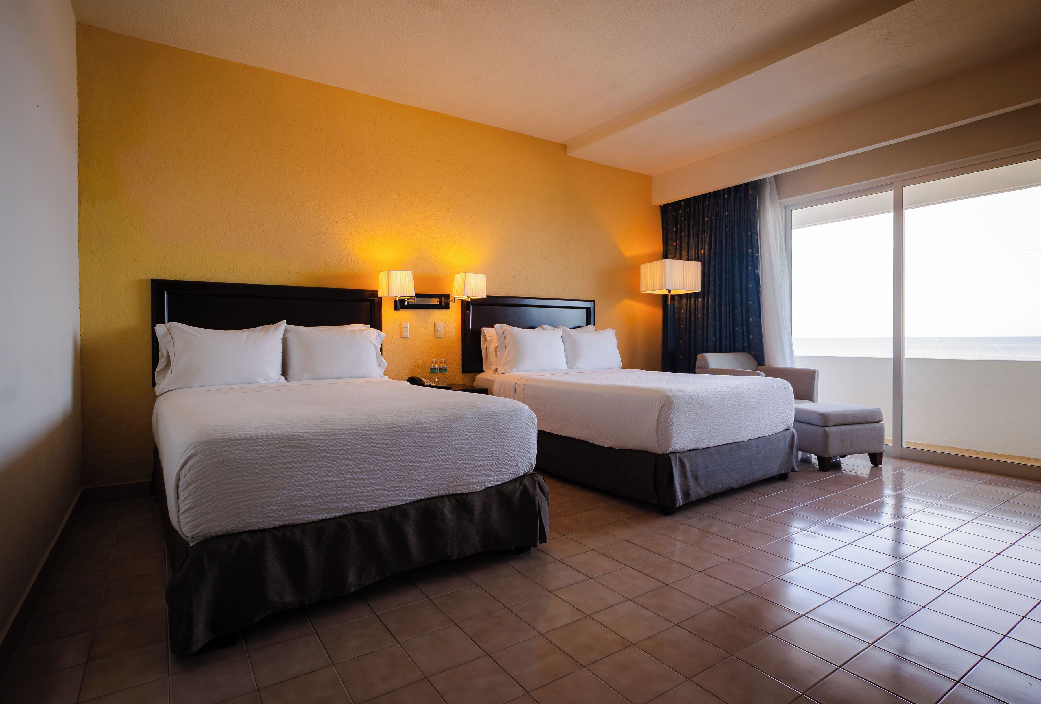 A hotel room with two beds, a small table, a chair, and a large window with a view.
