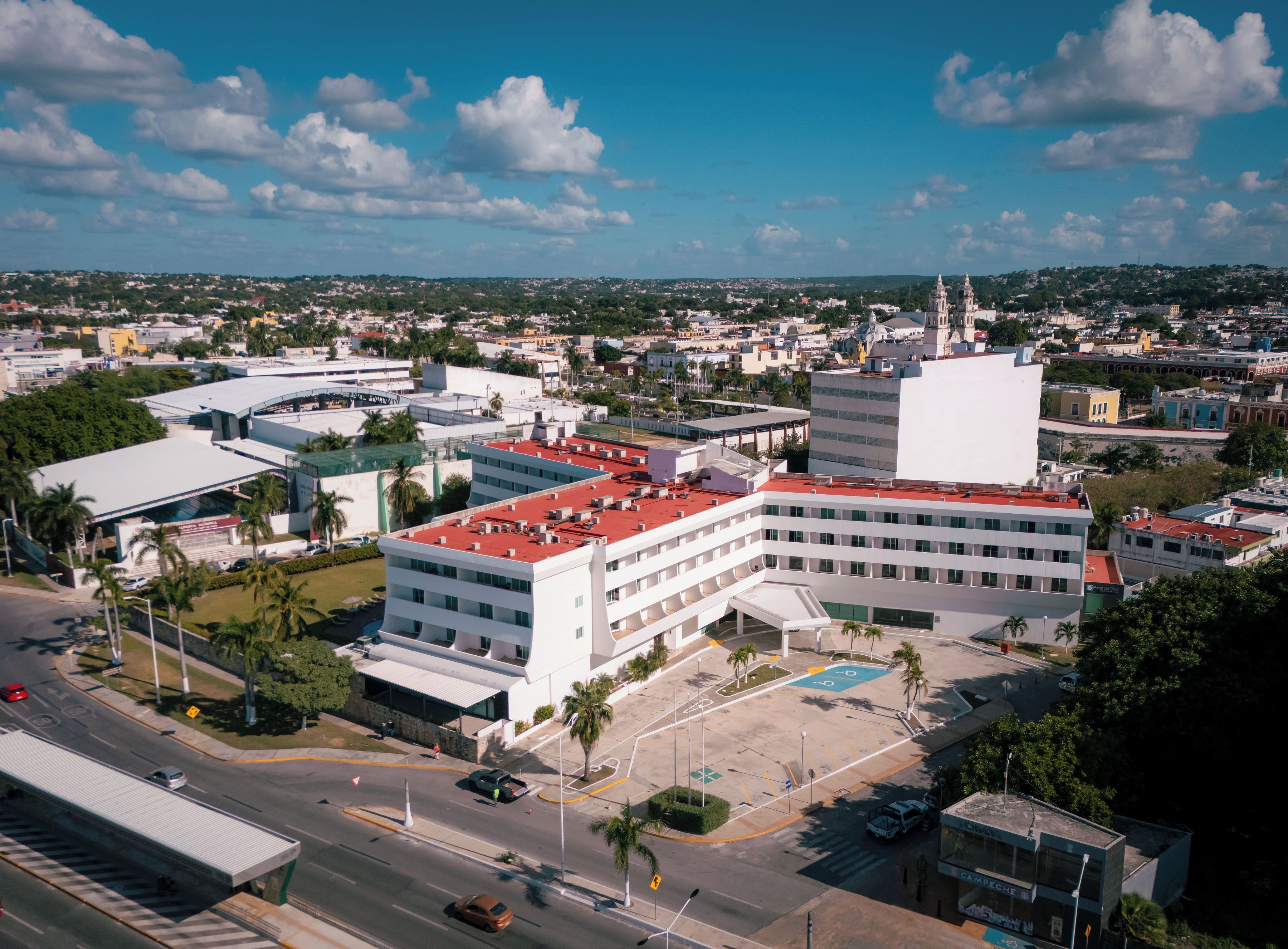 An aerial view of a cityscape with a prominent white building with a red roof, surrounded by smaller structures and greenery.