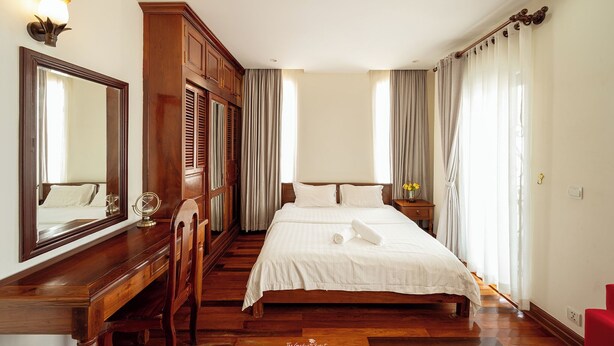 A neatly arranged bedroom with a wooden dresser, a large mirror, a bed with white linens, and a nightstand with a vase of flowers. - Cozy 1BR condo with hardwood floors and rooftop sunset garden in Phnom Penh (Phnom Penh)