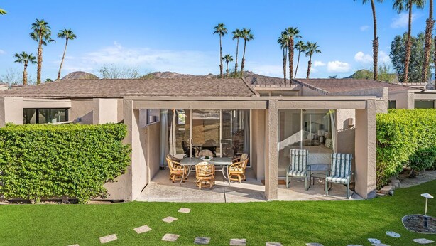 A modern house with a patio area featuring a dining table and chairs, surrounded by lush greenery and palm trees. - NEW! Mariposa Hideaway by GPM -Chic Ironwood CC Condo (Palm Desert)