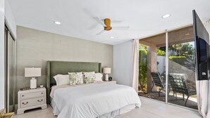 A modern bedroom with a large bed, two bedside tables with lamps, a ceiling fan, and a sliding glass door leading to an outdoor area with chairs. - NEW! Mariposa Hideaway by GPM -Chic Ironwood CC Condo (Palm Desert)
