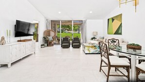 A spacious living room with a flat-screen TV, a dining area with a glass table, and a large window offering a view of a garden. - NEW! Mariposa Hideaway by GPM -Chic Ironwood CC Condo (Palm Desert)
