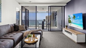 Un salon moderne avec un canapé gris, une table basse agrémentée d’objets décoratifs, un téléviseur posé sur un meuble en bois, et un balcon offrant une vue sur l’océan et les bâtiments.