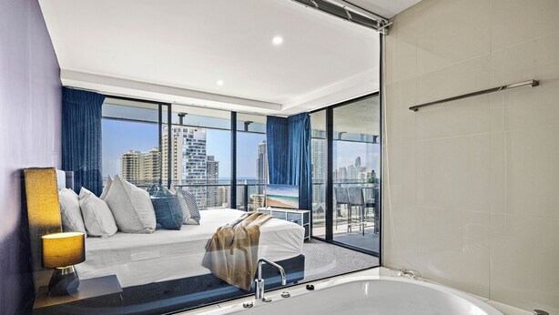 Une chambre d’hôtel moderne dotée d’une grande baignoire, d’un lit avec vue sur la ville et d’un balcon offrant un panorama sur le paysage urbain.