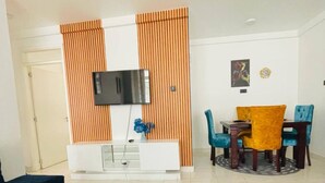 Una sala moderna con una mesa de comedor de madera oscura, un mueble blanco para entretenimiento con un televisor y un área de comedor con sillas azules y amarillas.