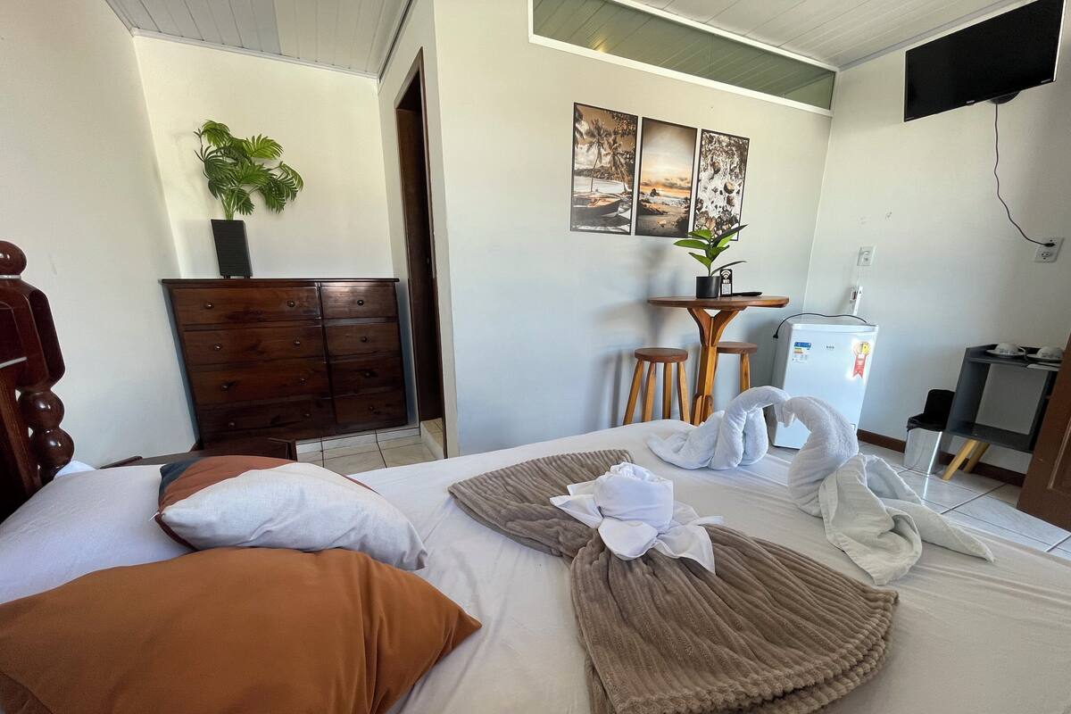 Ett hotellrum med en säng, en byrå, ett skrivbord med en stol, ett kylskåp och en tv som är monterad på väggen.
