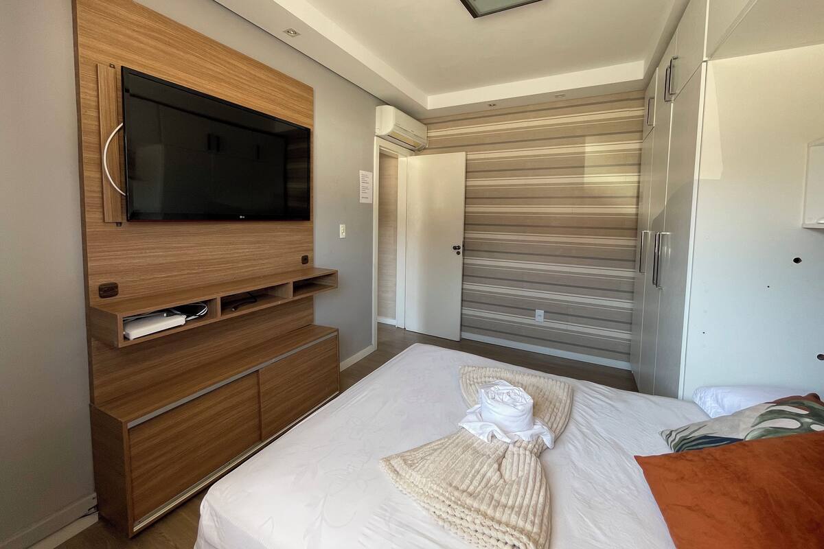 Ett modernt hotellrum med en stor platt-TV som är monterad på en träpanel, en skjutdörr till garderoben och en snyggt bäddad säng med en handduk och ett täcke.