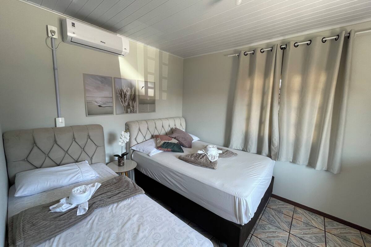 Ett hotellrum med två sängar, en takfläkt, en luftkonditioneringsanläggning, ett litet bord med en vas med blommor och ett fönster med gardiner.