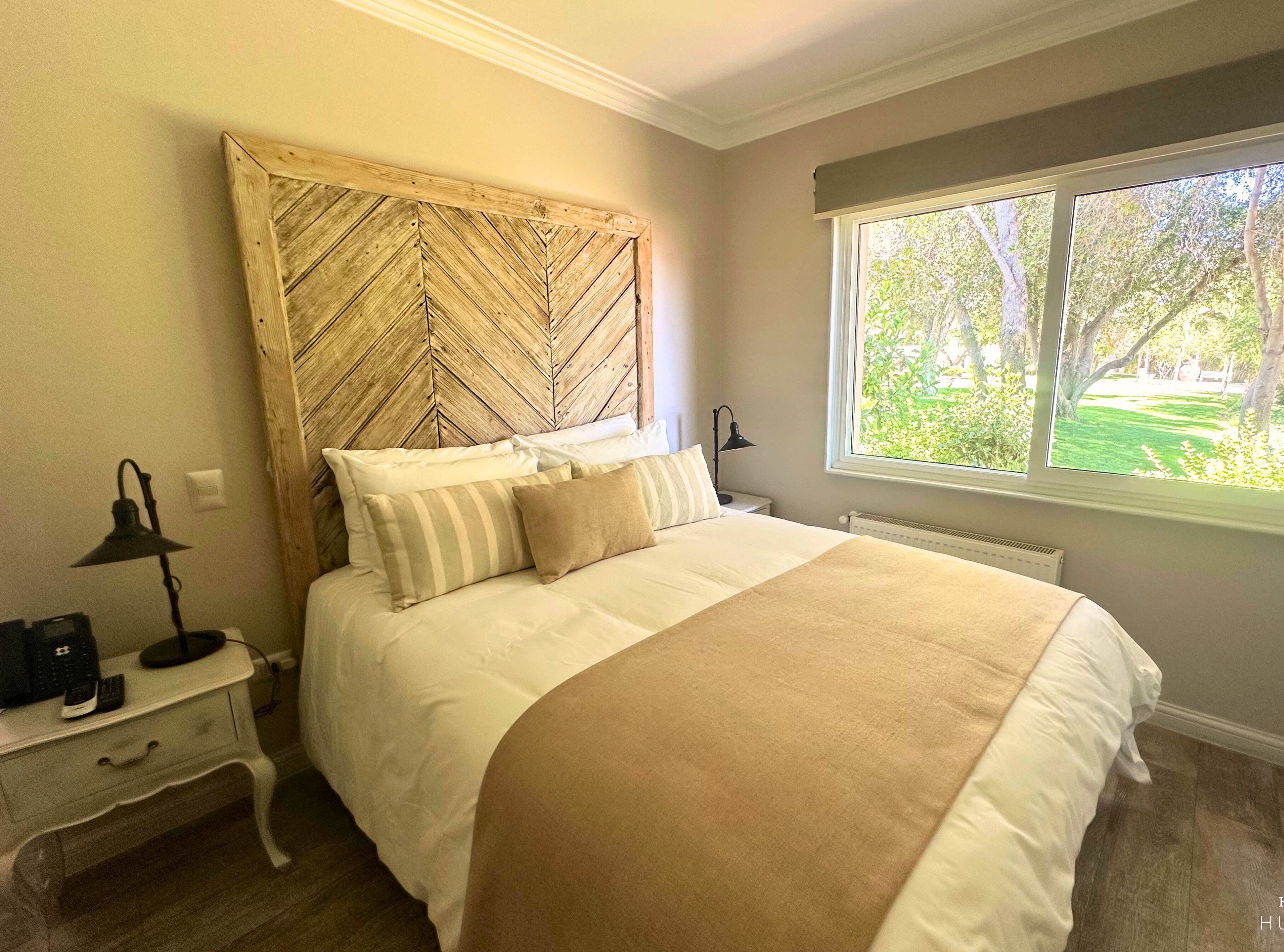 A bedroom with a bed, bedside tables, a window with a view of trees, and a wooden headboard.