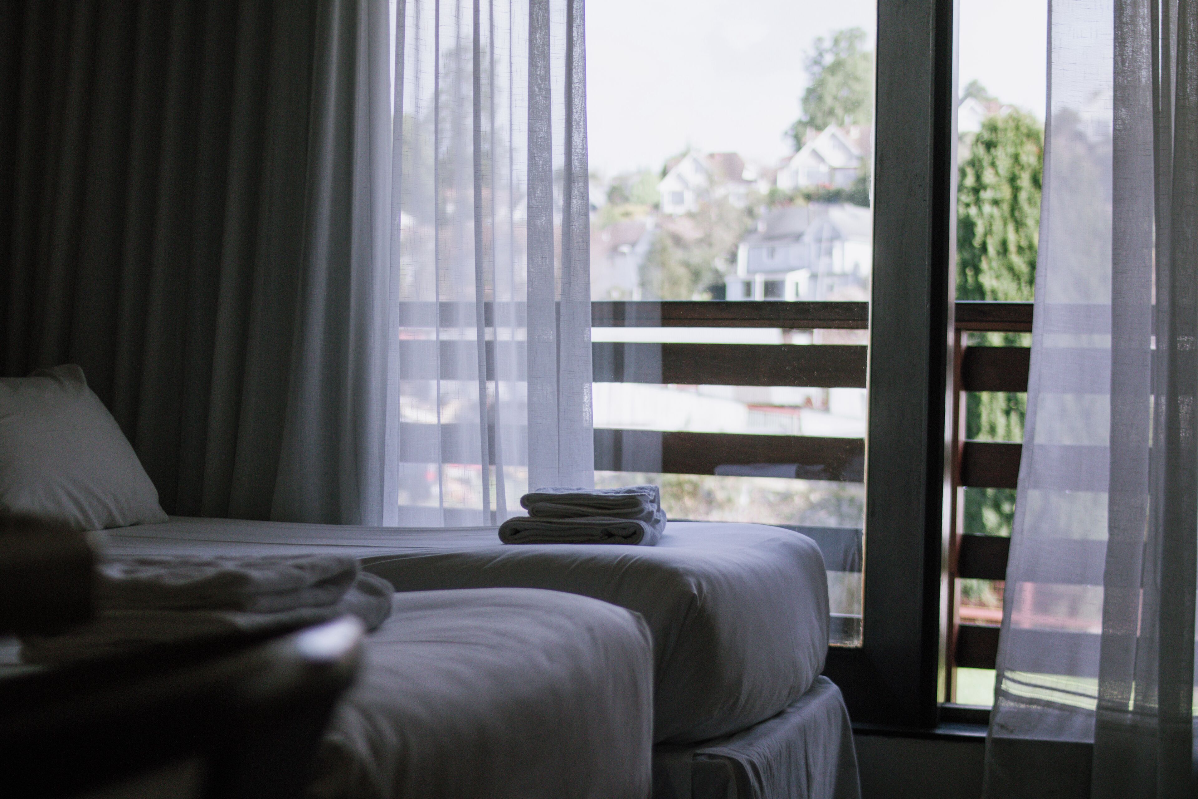 Habitación de hotel con dos camas, una ventana con cortinas traslúcidas y vistas a casas.