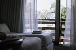 Habitación de hotel con dos camas, una ventana con cortinas traslúcidas y vistas a casas.