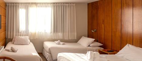 A hotel room with two single beds, a wooden wardrobe, a small table, and a window with sheer curtains.