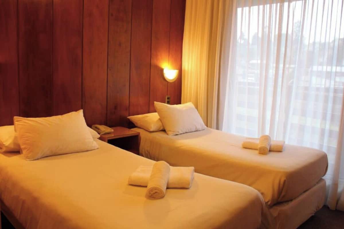 Two single beds with white linens and pillows, a wooden wall, a nightstand with a phone, and a window with sheer curtains.