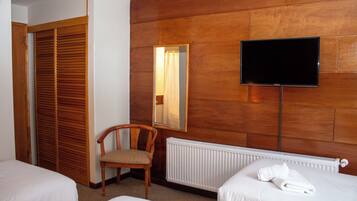 A hotel room with a wooden wall panel, a television mounted on the wall, a chair, a radiator, and two beds.
