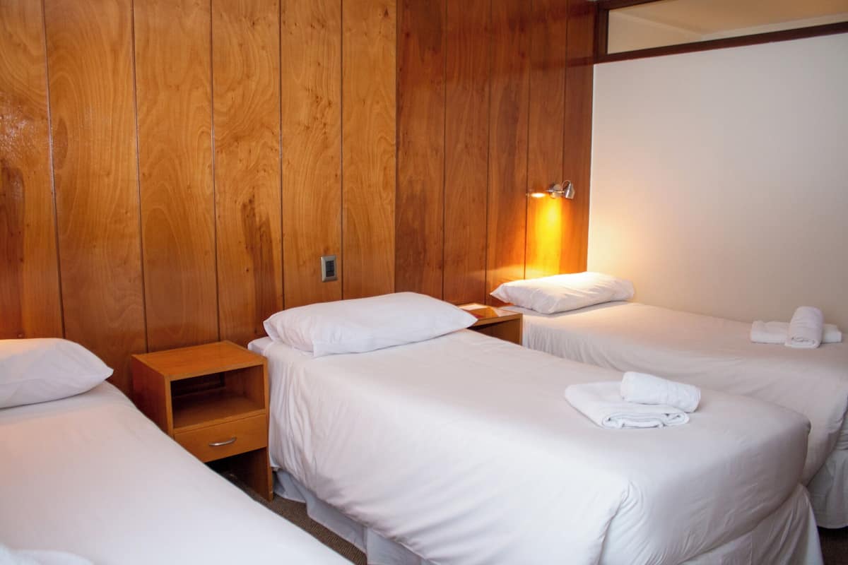 A hotel room with wooden panel walls, two single beds with white linens, a nightstand with a lamp, and a small wooden shelf.