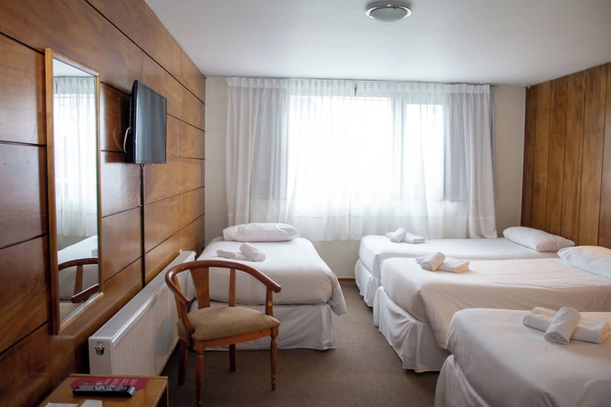 A hotel room with three beds, a wooden chair, a desk with a remote control, and a large mirror on the wall.