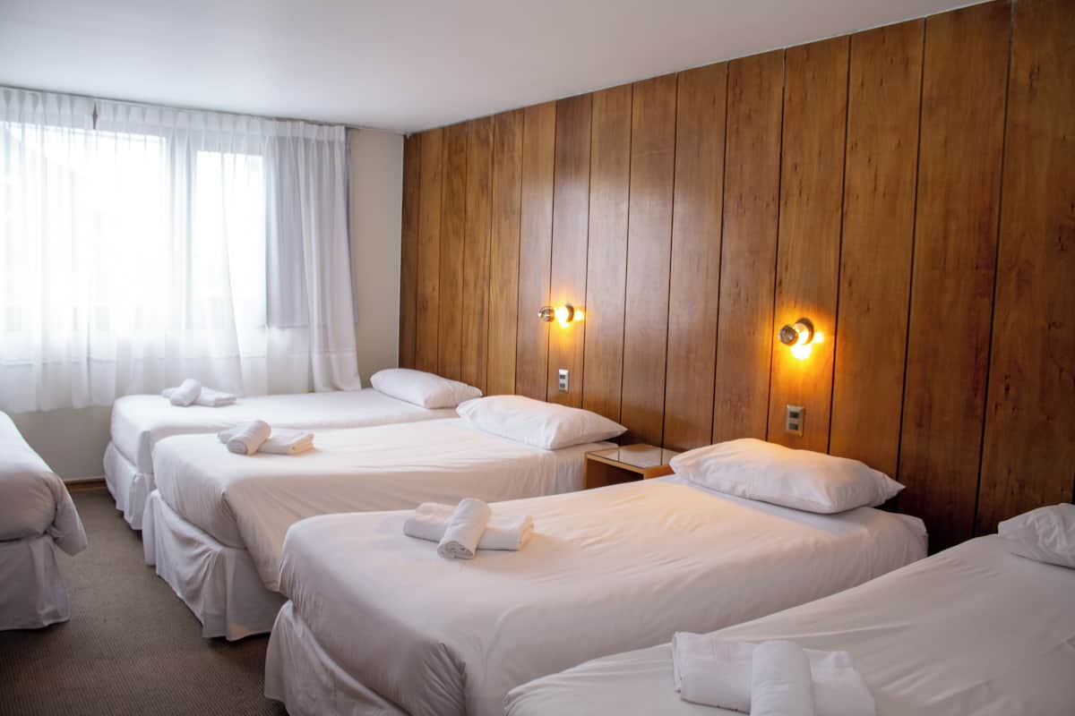 A hotel room with three single beds, each with a pillow and a rolled towel. There are two wall-mounted lights on the wooden panel wall.