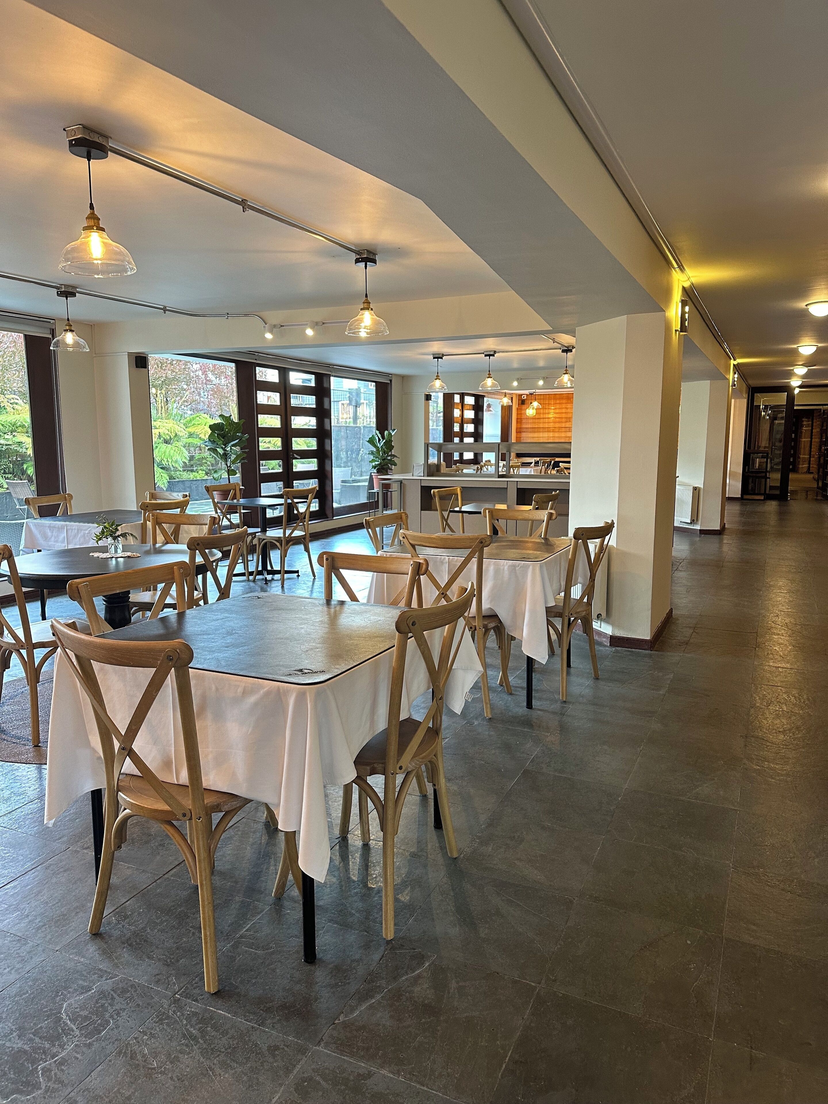 A spacious dining area with multiple tables and chairs, large windows offering a view of greenery, and pendant lighting.