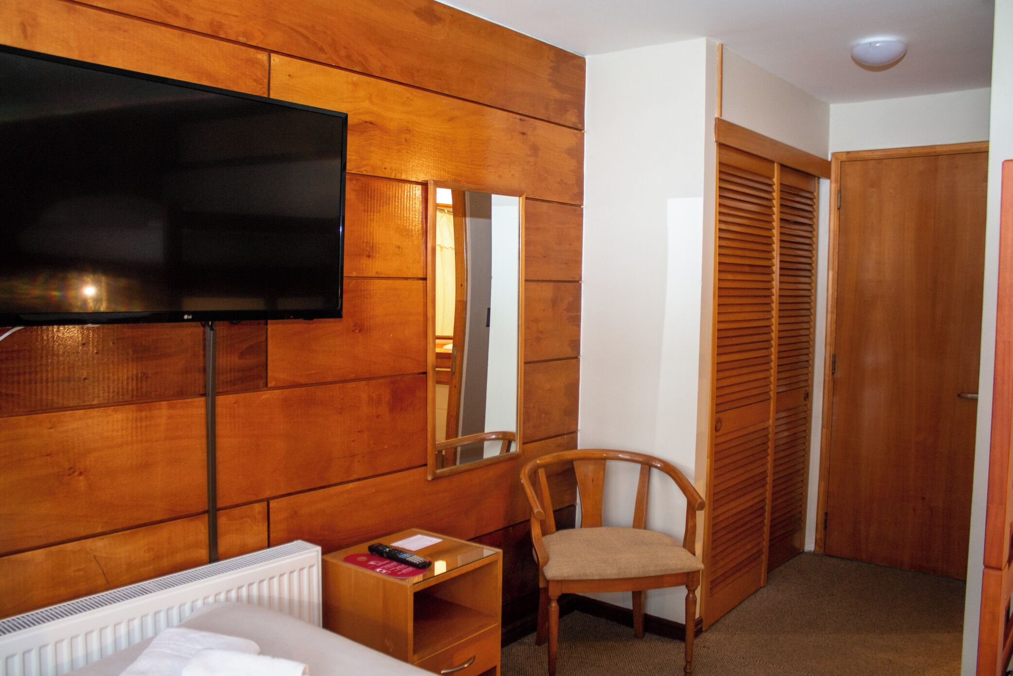 A hotel room with a wooden panel wall, a flat-screen TV, a wooden chair, a small table with a remote control, and a closet with sliding doors.