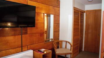A hotel room with a wooden panel wall, a flat-screen TV, a wooden chair, a small table with a remote control, and a closet with sliding doors.