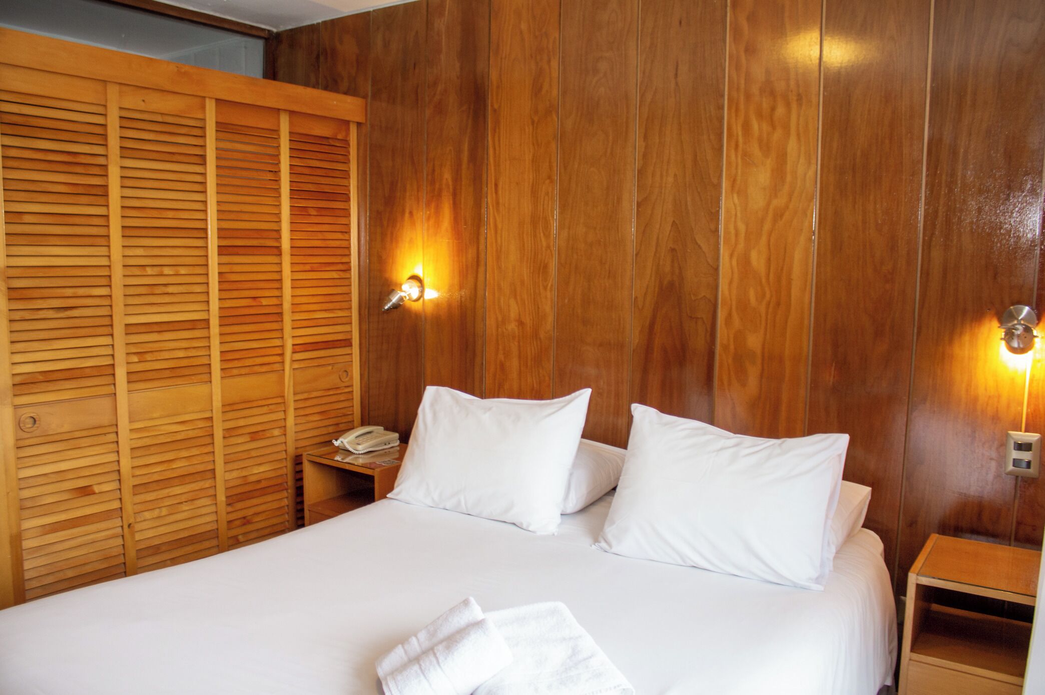 A hotel room with a wooden panel wall, a bed with white bedding, two bedside tables, and a wall-mounted light fixture.