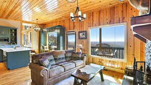 L’intérieur chaleureux d’un chalet avec un plafond en bois, un canapé agrémenté de coussins à motifs, une table à manger et une grande fenêtre donnant sur le paysage extérieur.