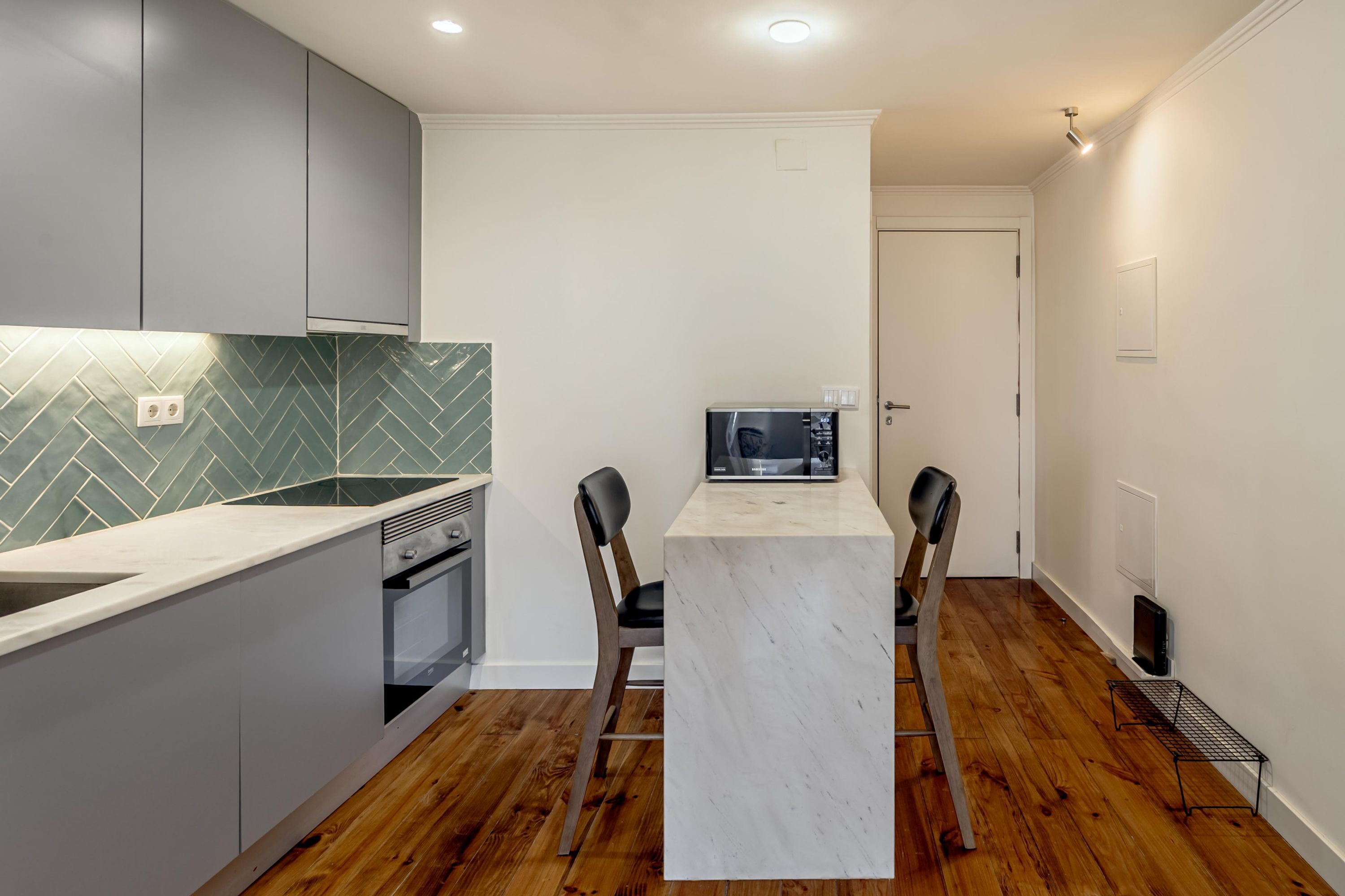 Uma cozinha compacta com painel de azulejos em padrão herringbone, micro-ondas e bancada de mármore.