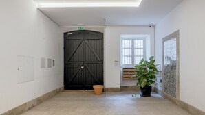 A hallway with a large black door, a window with blinds, a potted plant, and a tiled floor. - Bairro Alto Garden With A View by Homing (Lisbon)