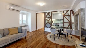 Una sala de estar moderna con piso de madera, un sofá gris, una mesa de comedor de vidrio con sillas y una cocina al fondo.