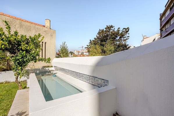 Una piscina con bordo piastrellato, circondata da un muro bianco e vegetazione.