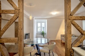 A dining area with a glass table surrounded by chairs, wooden beams, a potted plant, and a window with a view of buildings. - Bairro Alto Garden With A View II by Homing (Lisbon)