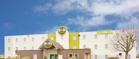 A B&B hotel with a distinct yellow and white facade, a green entrance, and a visible parking area.