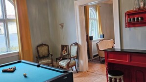 A billiards table with balls, a pool cue, and a cue rack. There are two chairs with cushioned seats and wooden frames. A red bar counter with stools is visible. The room has wooden flooring and a decorative ceiling with a chandelier.
