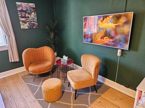 Une pièce comprenant deux fauteuils, un petit pouf, une plante en pot et un tableau accroché au mur.