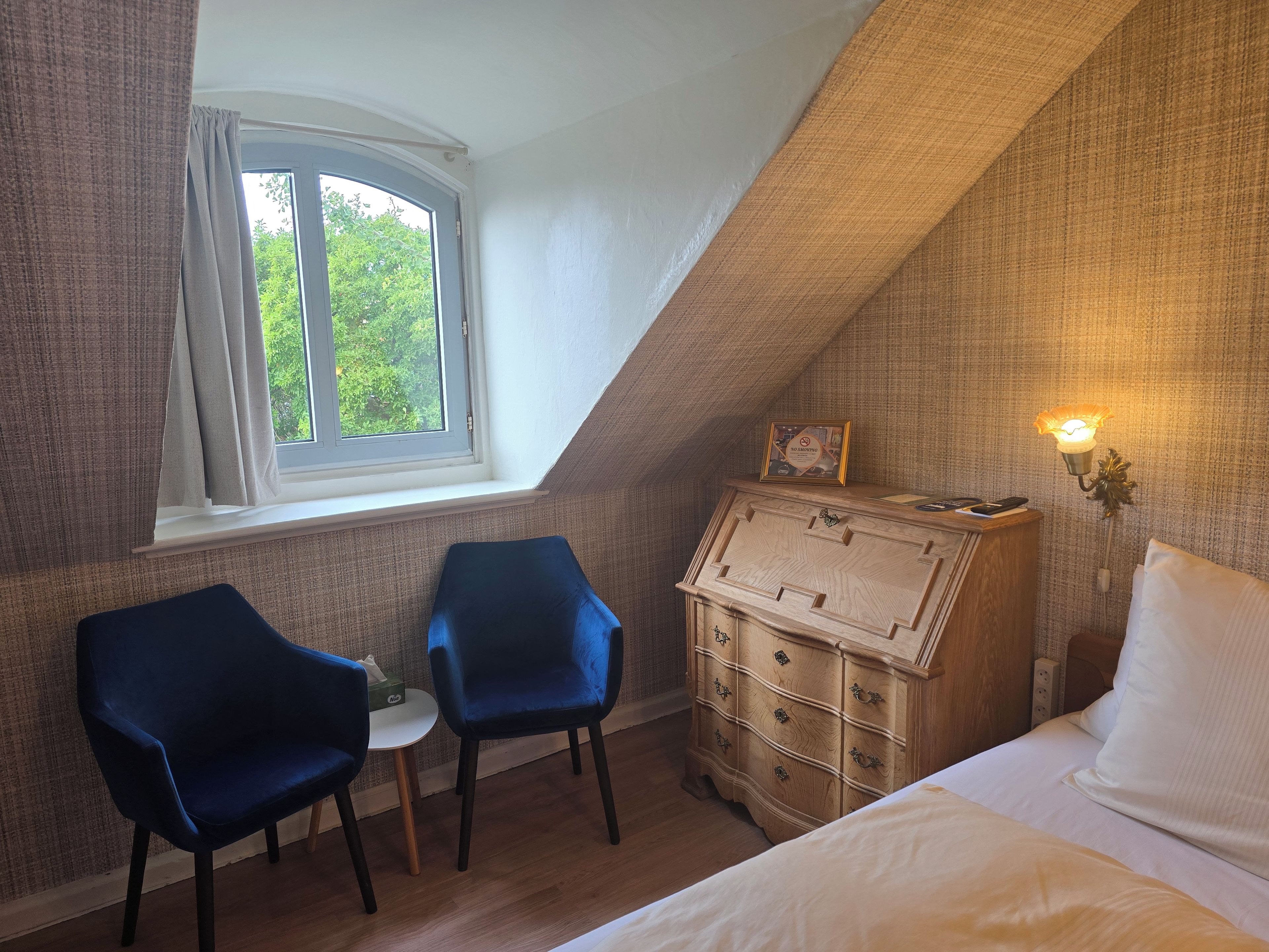 A bedroom with a bed, two chairs, a dresser, a window, and a wall-mounted light fixture.