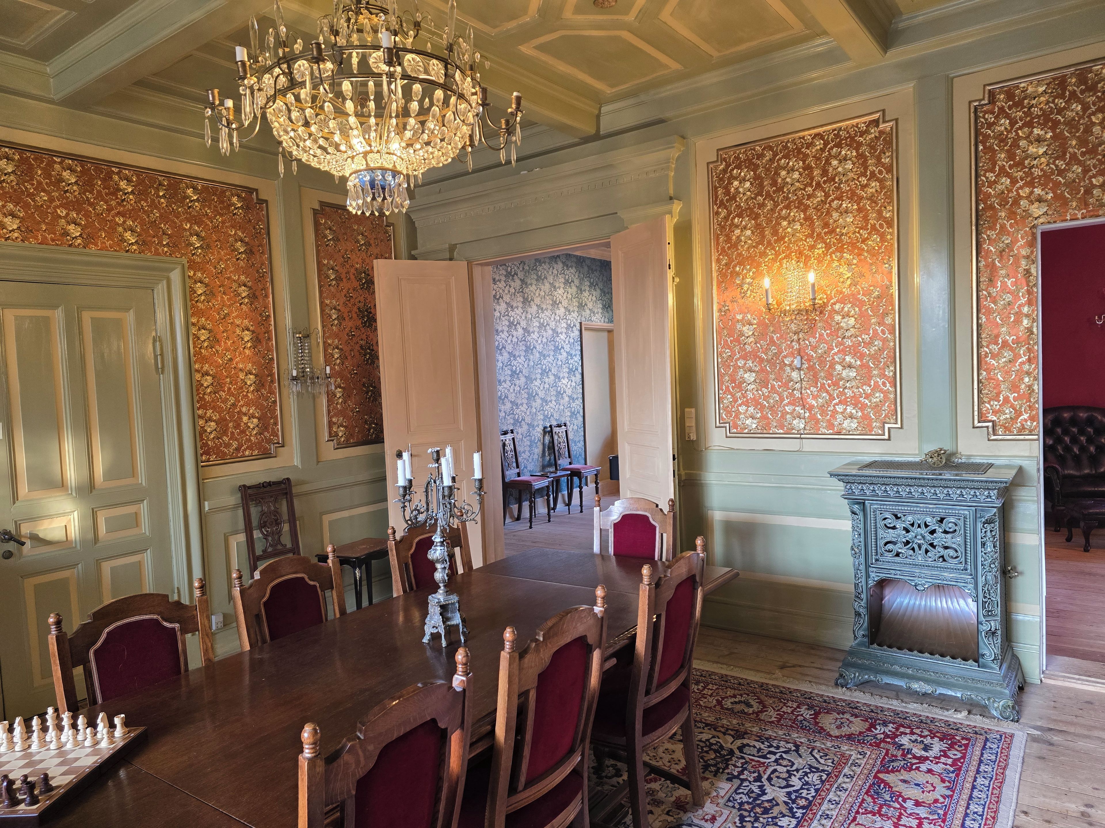 Comedor formal con una gran mesa de madera, sillas ornamentadas, un candelabro y papel tapiz estampado.