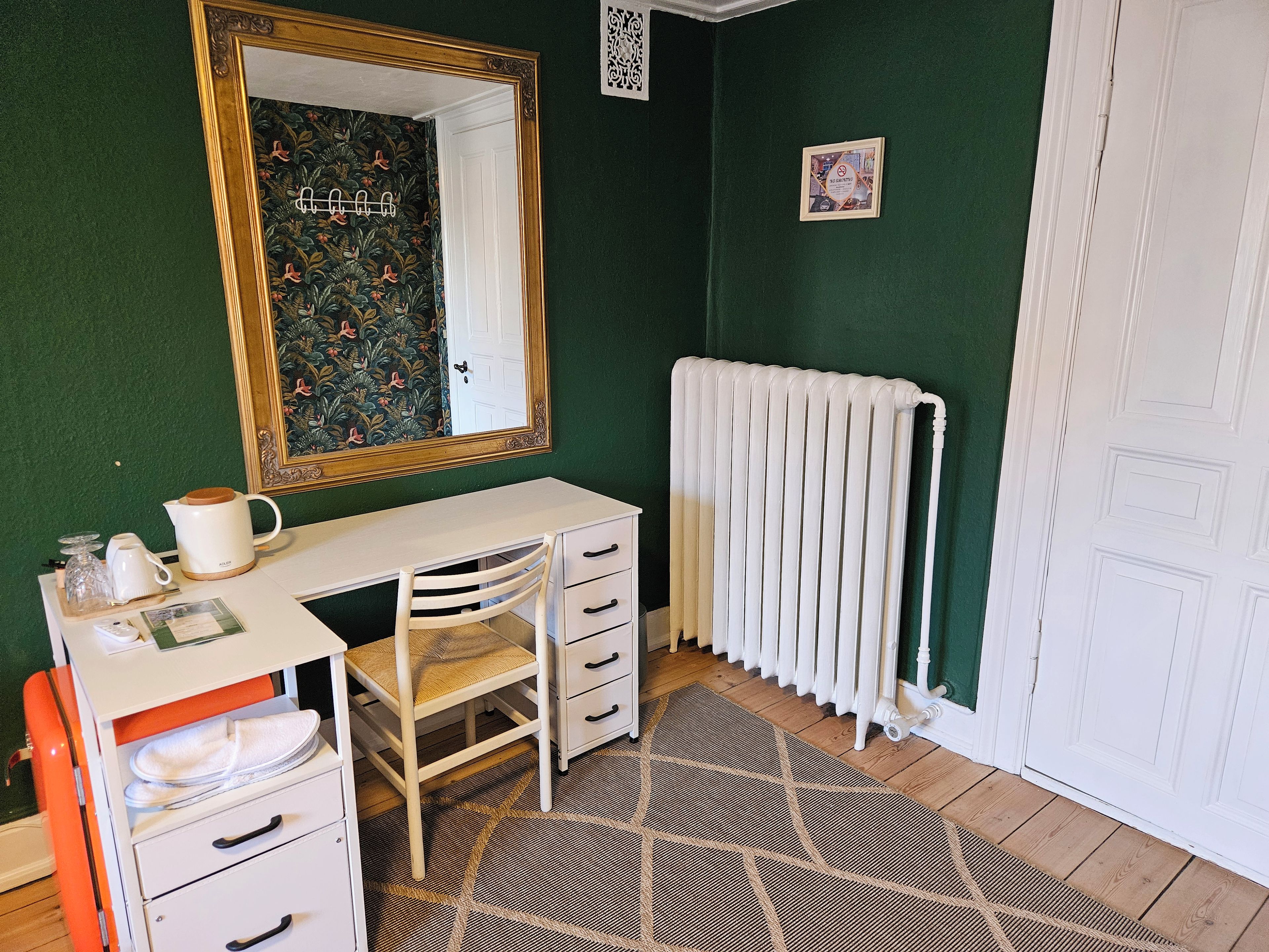 A room with a white desk, a chair, a radiator, and a mirror on the wall.