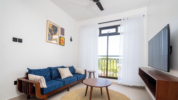 Ein modernes Wohnzimmer mit einem blauen Sofa, einem Holztisch, einem Deckenventilator, einem Fernseher und einem Balkon mit weißen Vorhängen.