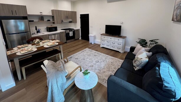 Ett modernt kök med rostfria vitvaror, en matplats med ett bord för två personer och ett vardagsrum med en svart soffa och ett litet runt bord.