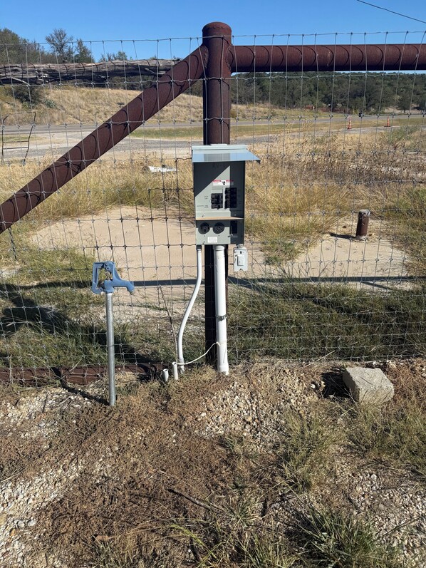 A utility box with a metal pole, surrounded by a chain-link fence, with a large metal pipe in the background.