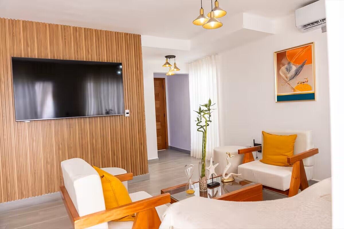 Een moderne woonkamer met een flatscreen televisie, een wand van houten panelen, witte banken met gele kussens en een schilderij aan de muur.