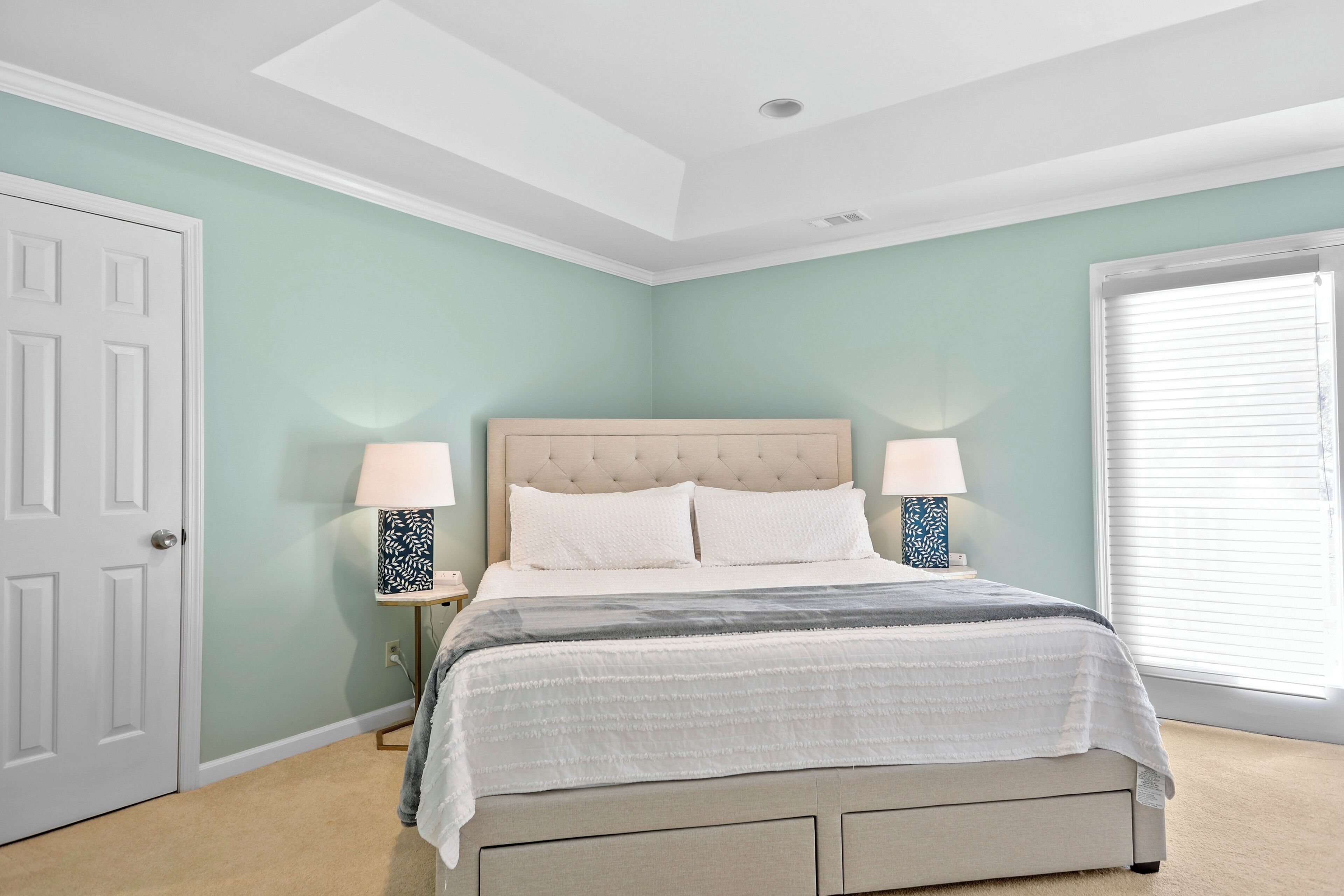 A bedroom with a bed, two bedside lamps, a window with blinds, and a door.