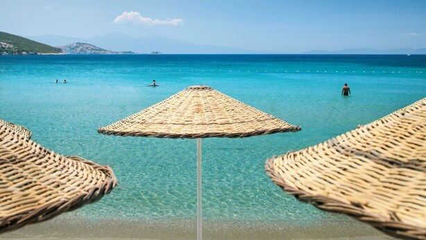 A beach with clear turquoise water, a straw umbrella, and a person standing in the water.