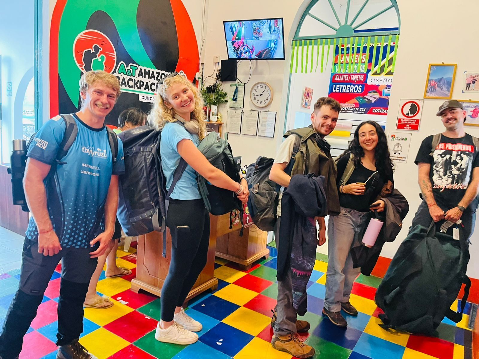 A group of people standing in a room with colorful checkered flooring, a large circular logo on the wall, and a television mounted on the wall.