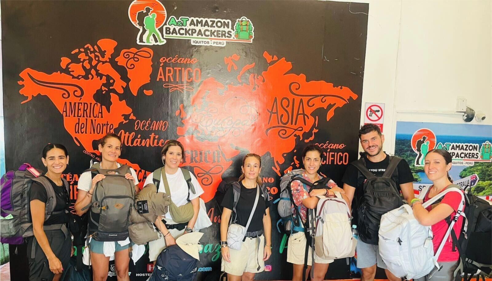 Un grupo de personas con mochilas frente a un telón de fondo con un mapa mundial y el texto 'A&T AMAZON BACKPACKERS' y 'IQUITOS - PERU'.