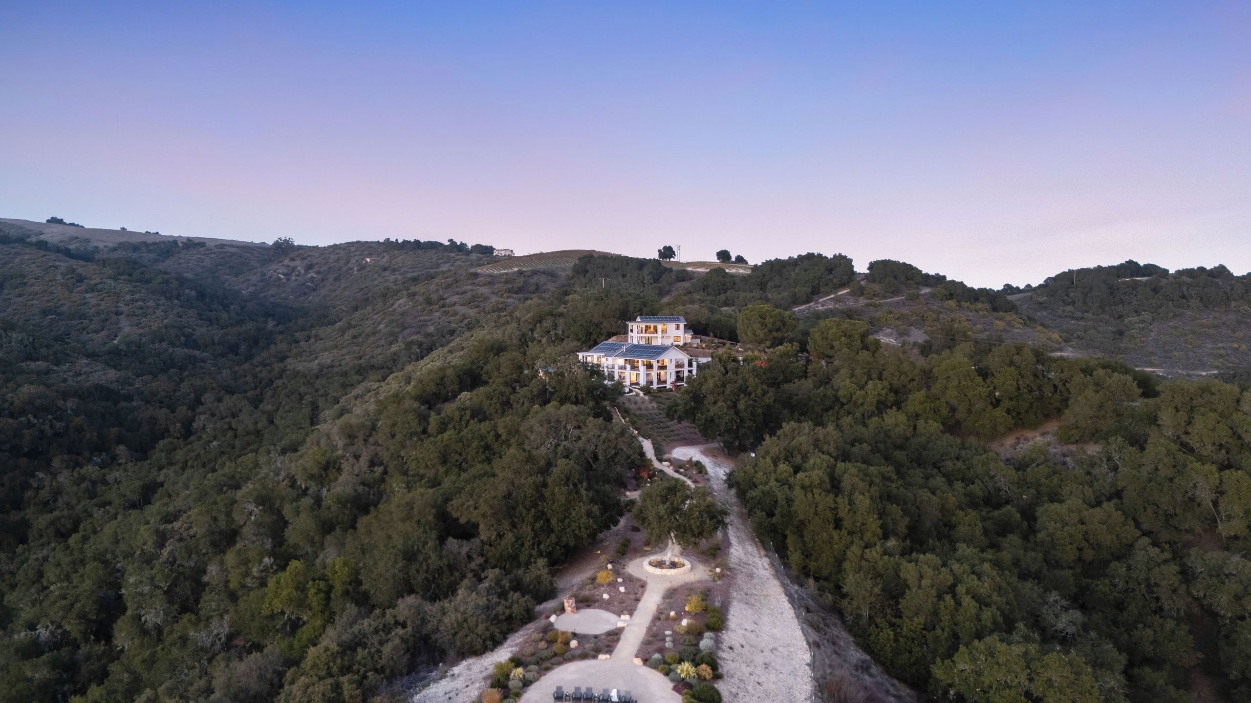 Una grande casa con un giardino e un vialetto che vi conduce, circondata da colline e alberi.