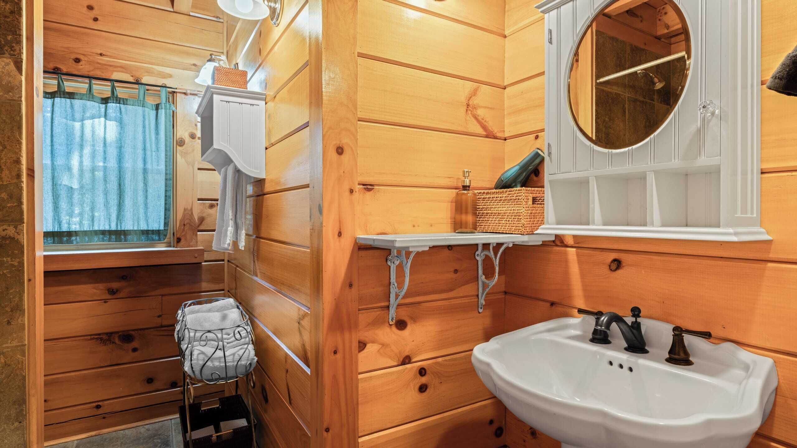 Baño de cabaña de madera con lavamanos, espejo y una pequeña ventana con cortinas.