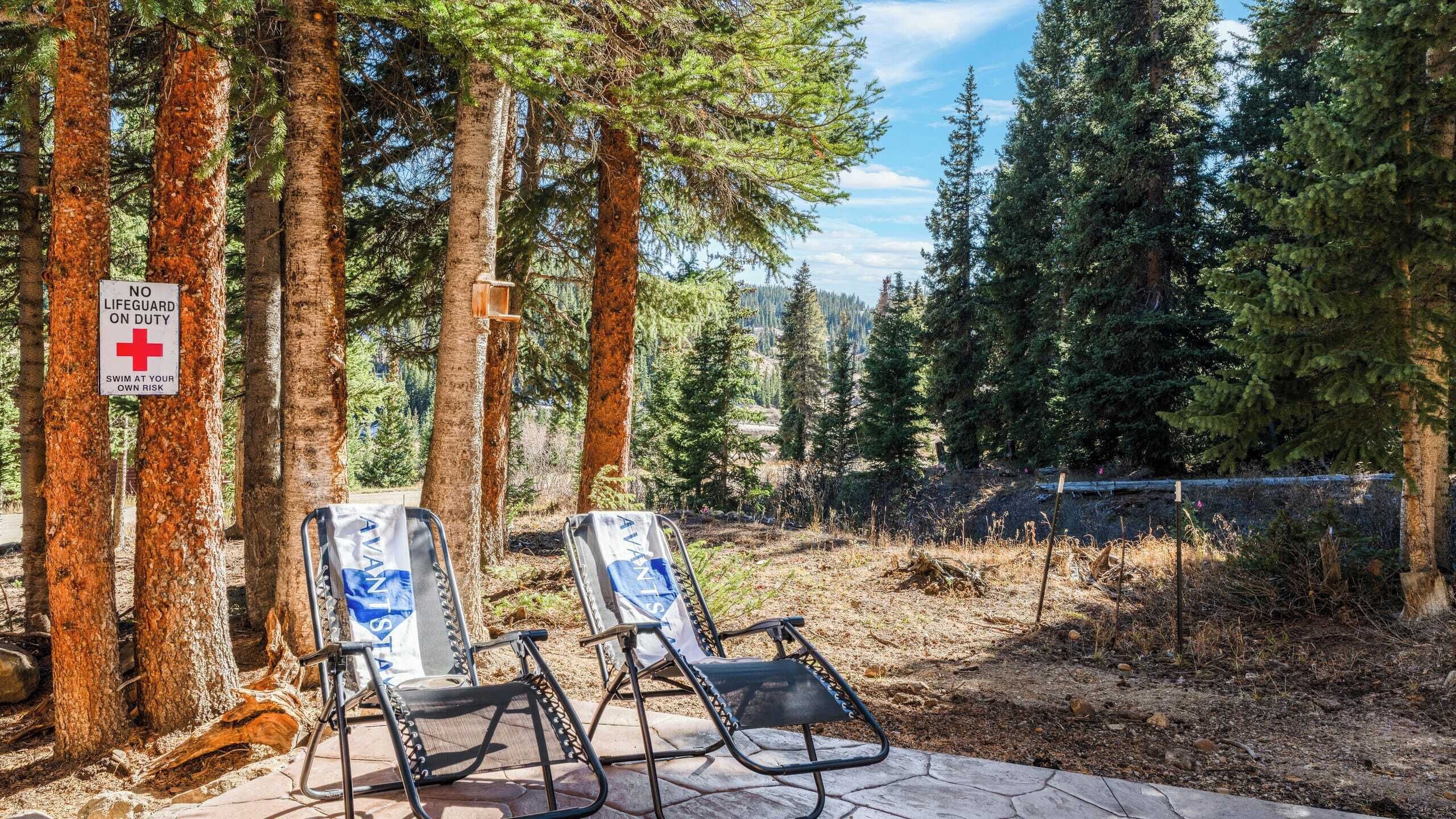 Two reclining chairs with a 'NO LIFEGUARD ON DUTY' sign, surrounded by tall trees and a clear sky.