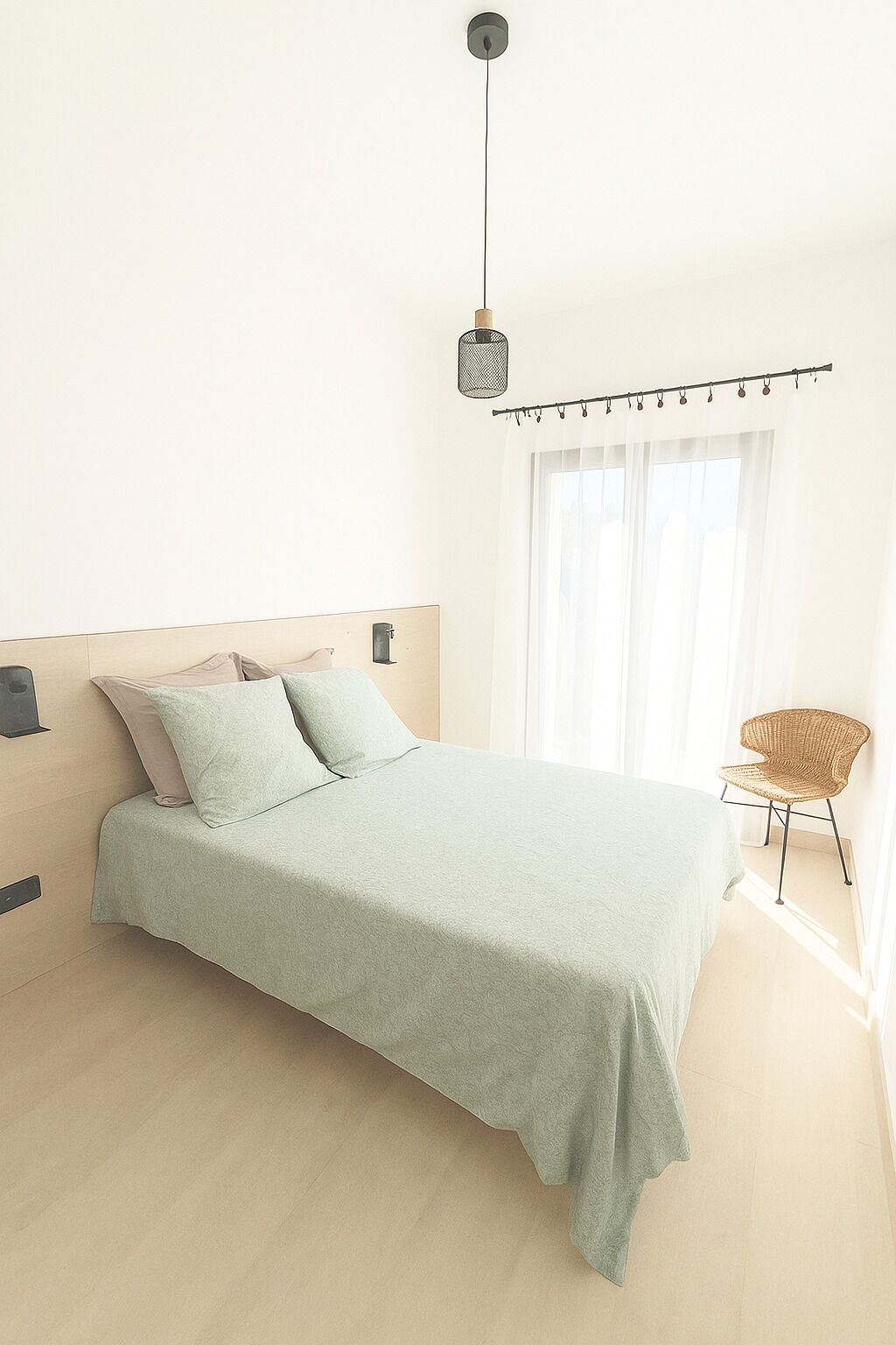 Un dormitorio con cama, lámpara colgante, silla y ventana con cortinas.