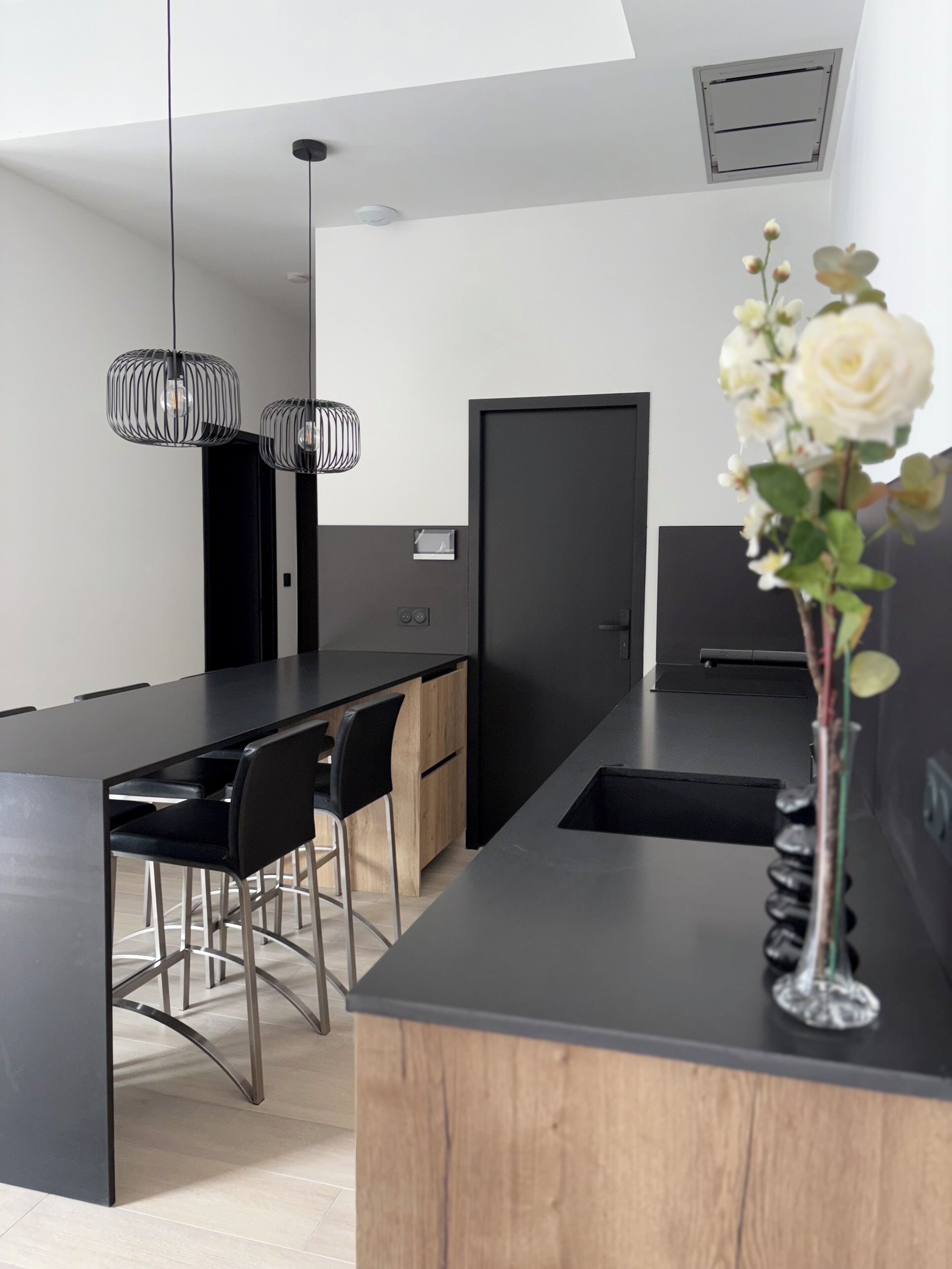 A modern kitchen with a central island, black bar stools, pendant lighting, and a vase with flowers.